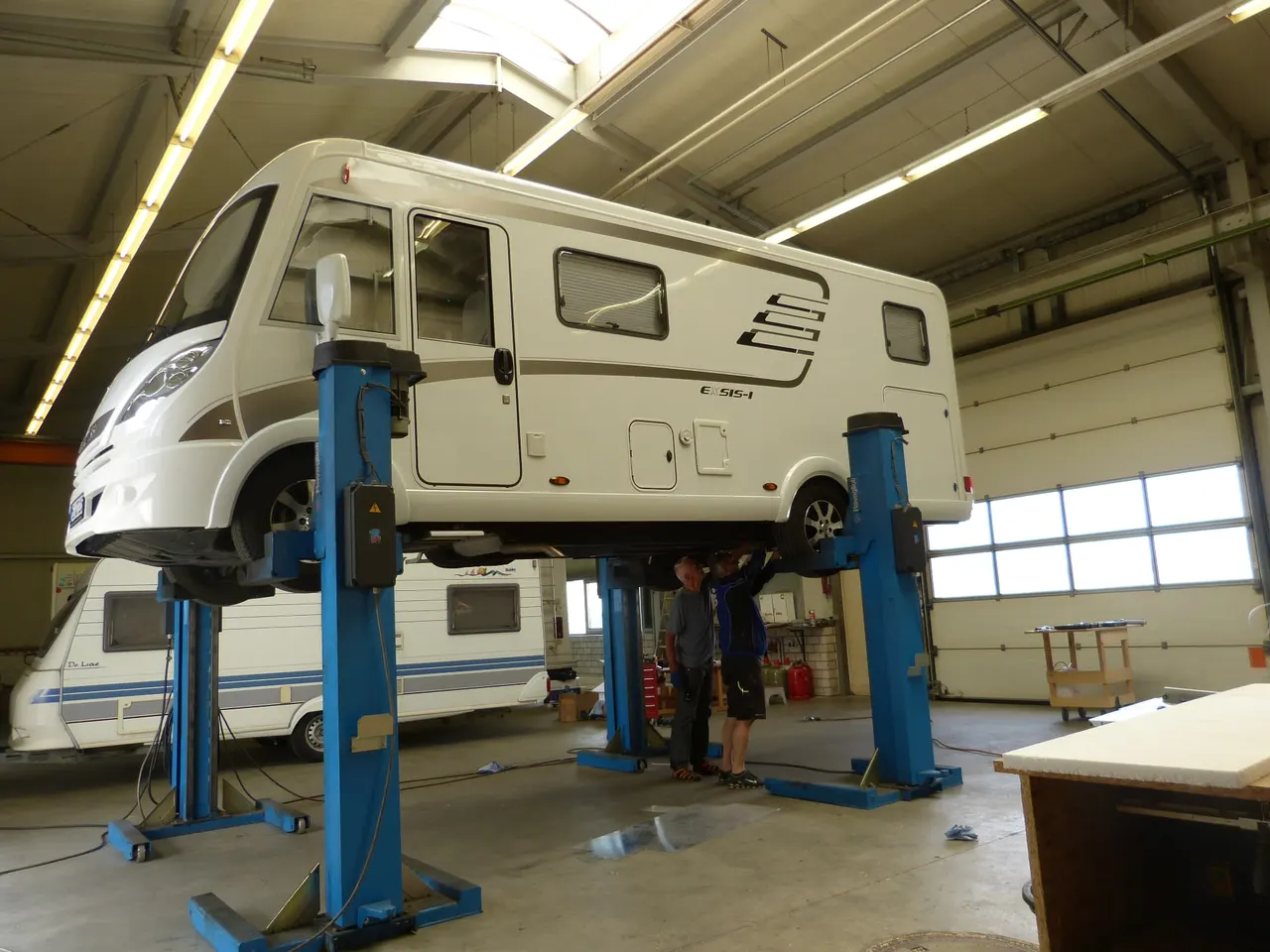 Wohnmobil auf blauen Hebebühnen in der Werkstatthalle, darunter zwei Personen bei der Arbeit am Unterboden.